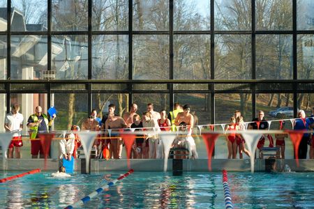 Blick entlang eines Schwimmbeckens: Im Wasser sind zwei Personen zu sehen, die um die Wette schwimmen. Am Beckenrand stehen Dutzende Betreuer*innen und Kinder zum Anfeuern.
